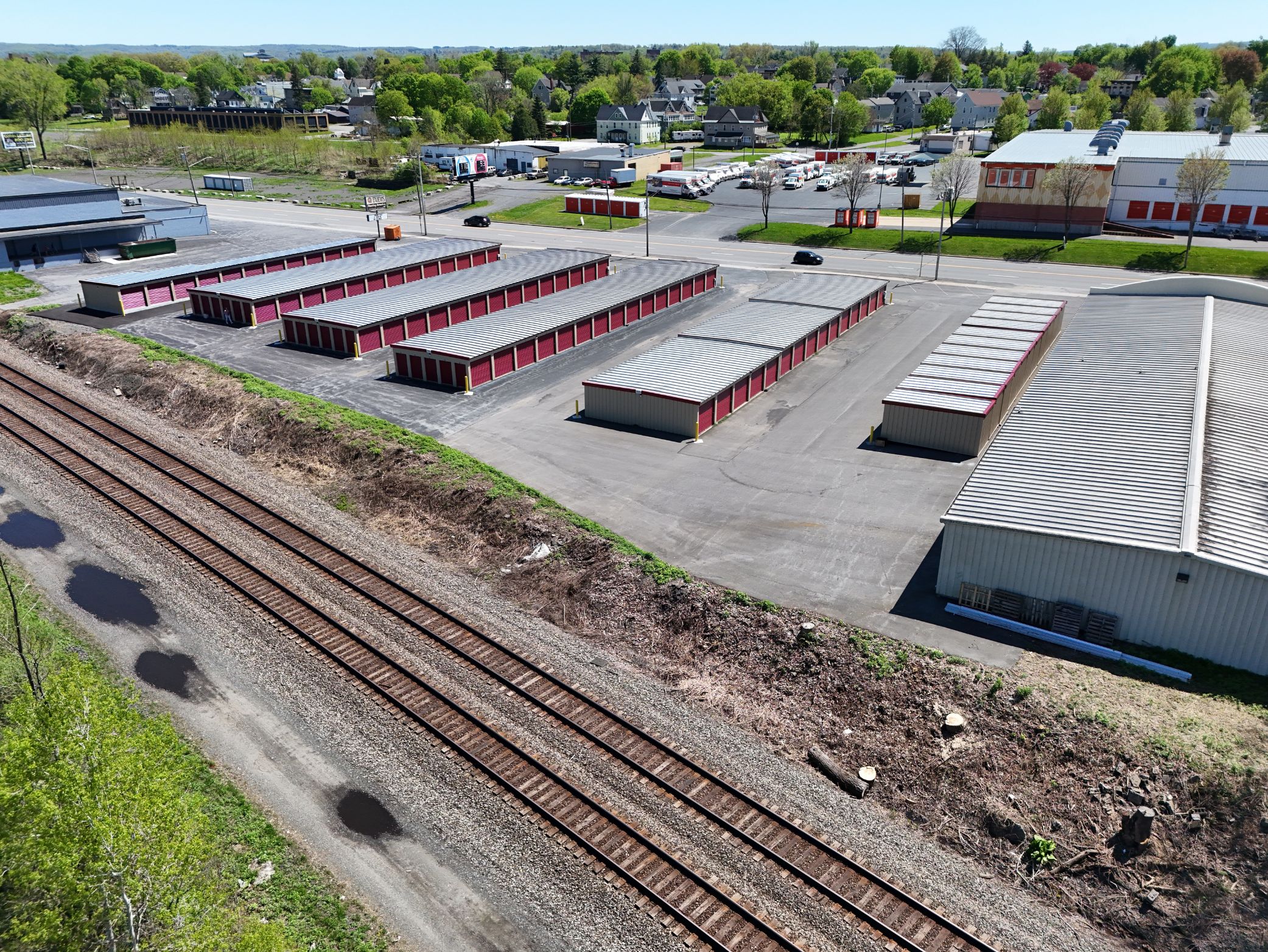 Aerial view of self storage units with red doors, paved driveways, nearby railroad tracks, and surrounding commercial buildings.